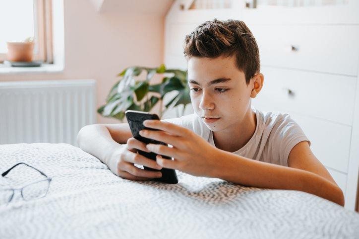Young teenage boy on his phone, propping up his arms on the mattress of his bed.