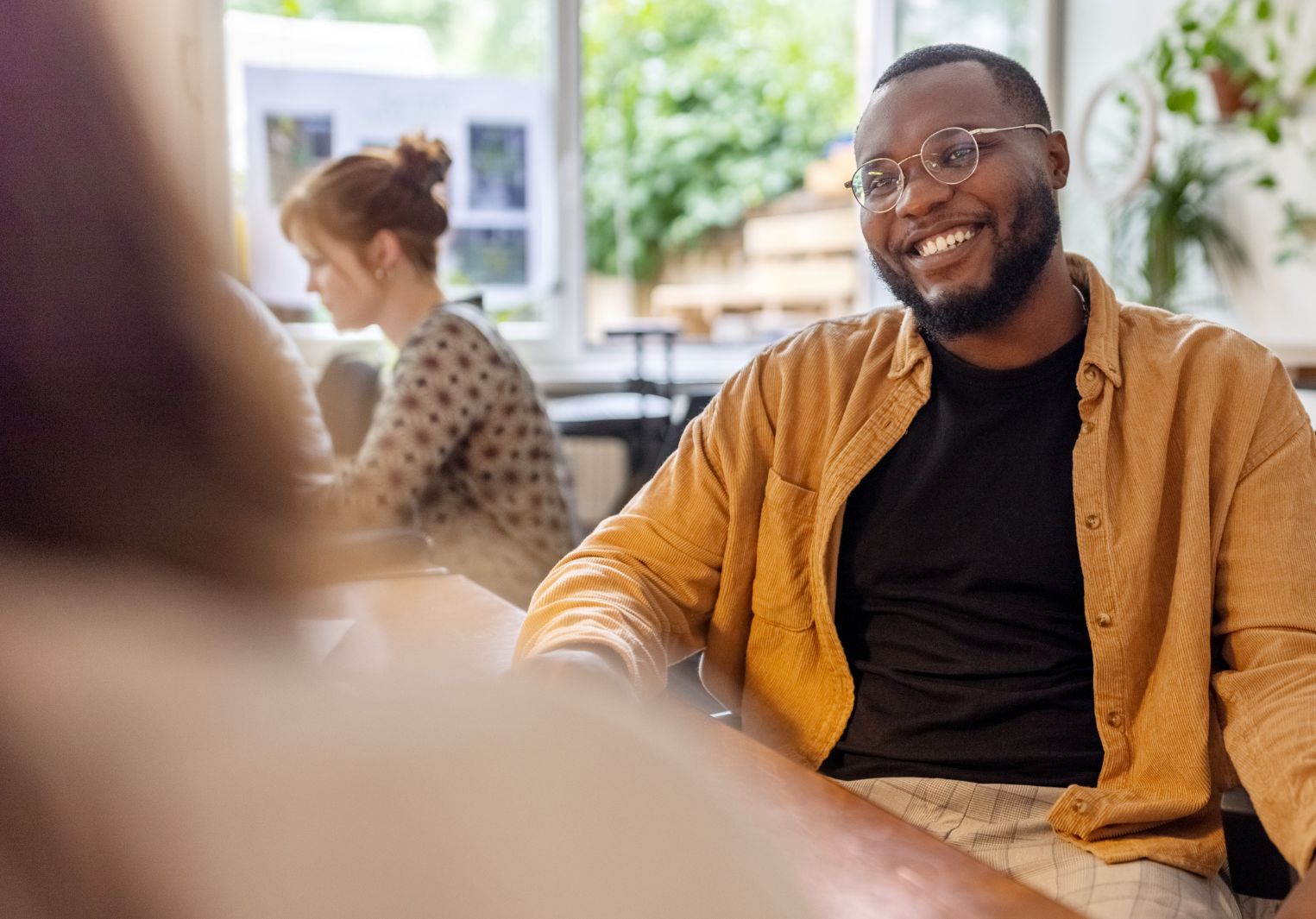 A young man in an office, wearing a warm yellow shirt and glasses smiling at someone sitting off-screen
