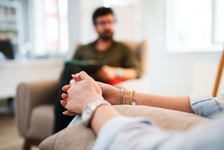 Close up of a woman's hands folded on her knees with a male therapist sitting in the background, out of focus