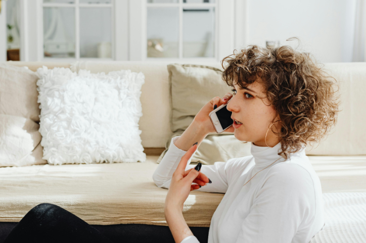 Young woman speaking on a mobile phone, sitting on the floor and leaning back against a white couch.