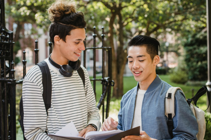 Two young men standing outside, holding a binder between them and talking about something on the page. 