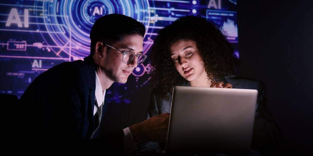Two people using a computer together. Behind them is a digital backdrop that says 'AI'.