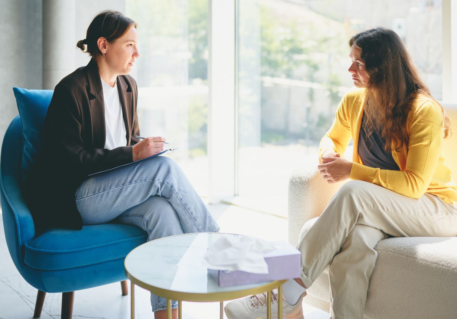 Two women sitting in a therapist's office, facing each other, mid conversation.