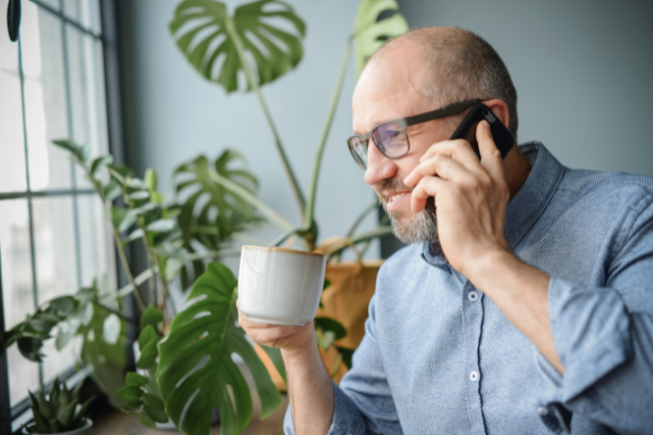 An older man on his phone, holding a coffee in the other hand and smiling.