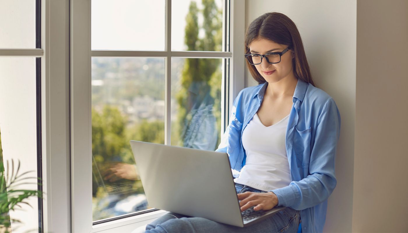 Young girl sits near window using her laptop.