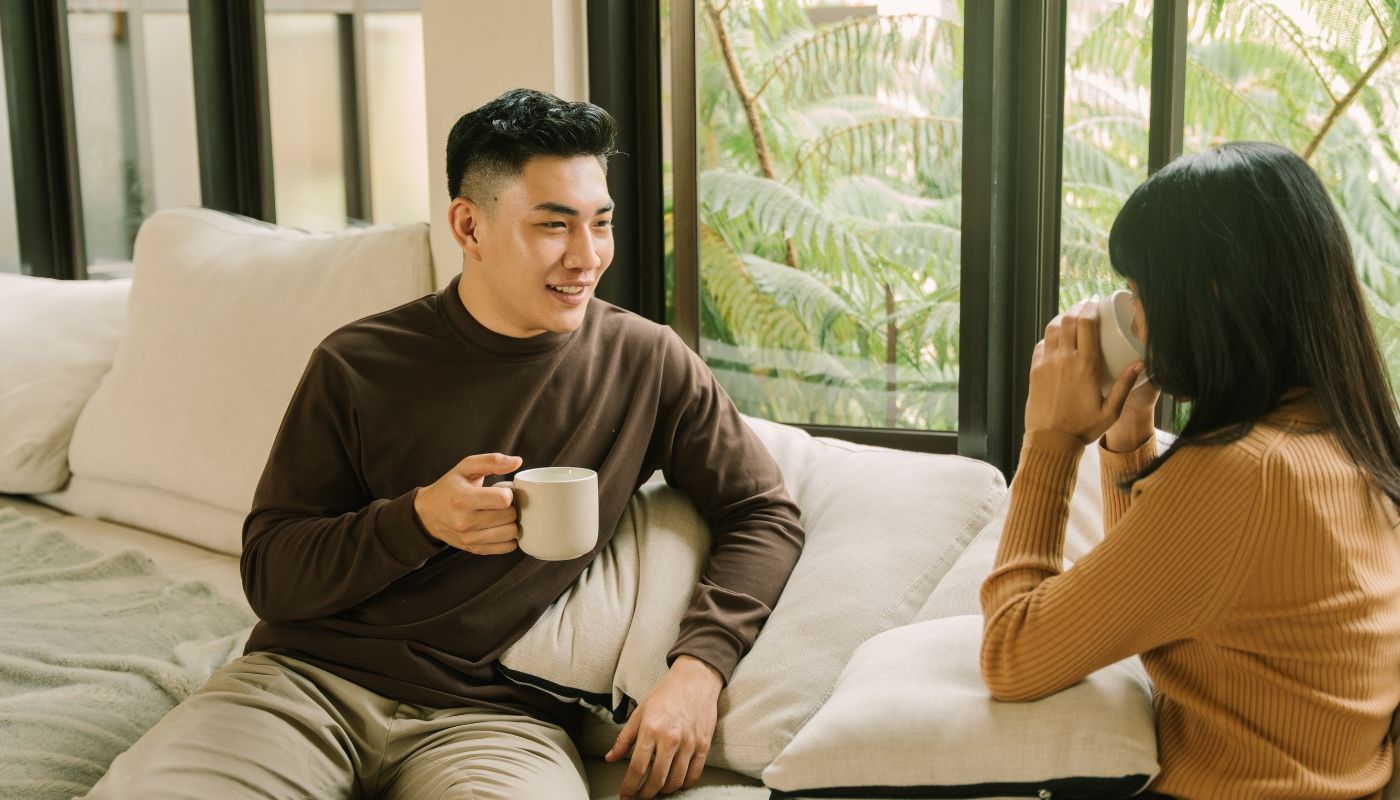 Man and woman sit talking on lounge while drinking coffee.