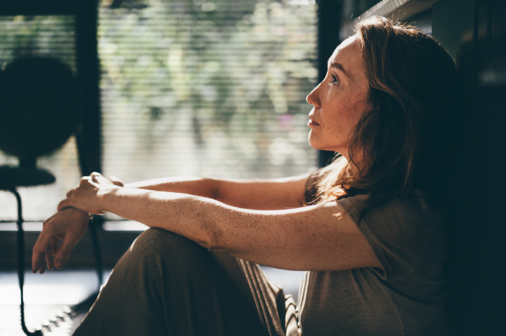 Side view of a woman sitting on the ground, resting her back against the wall and looking up sadly.