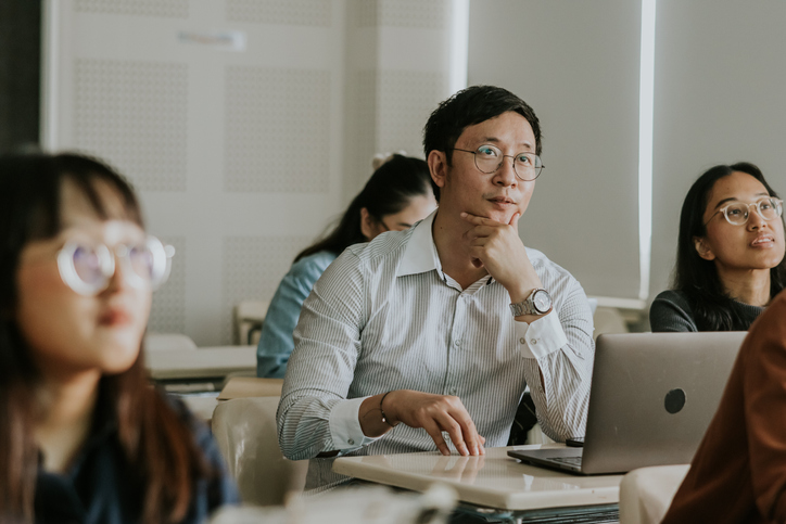 Asian man attending an in-person university class, paying attention to someone off camera.