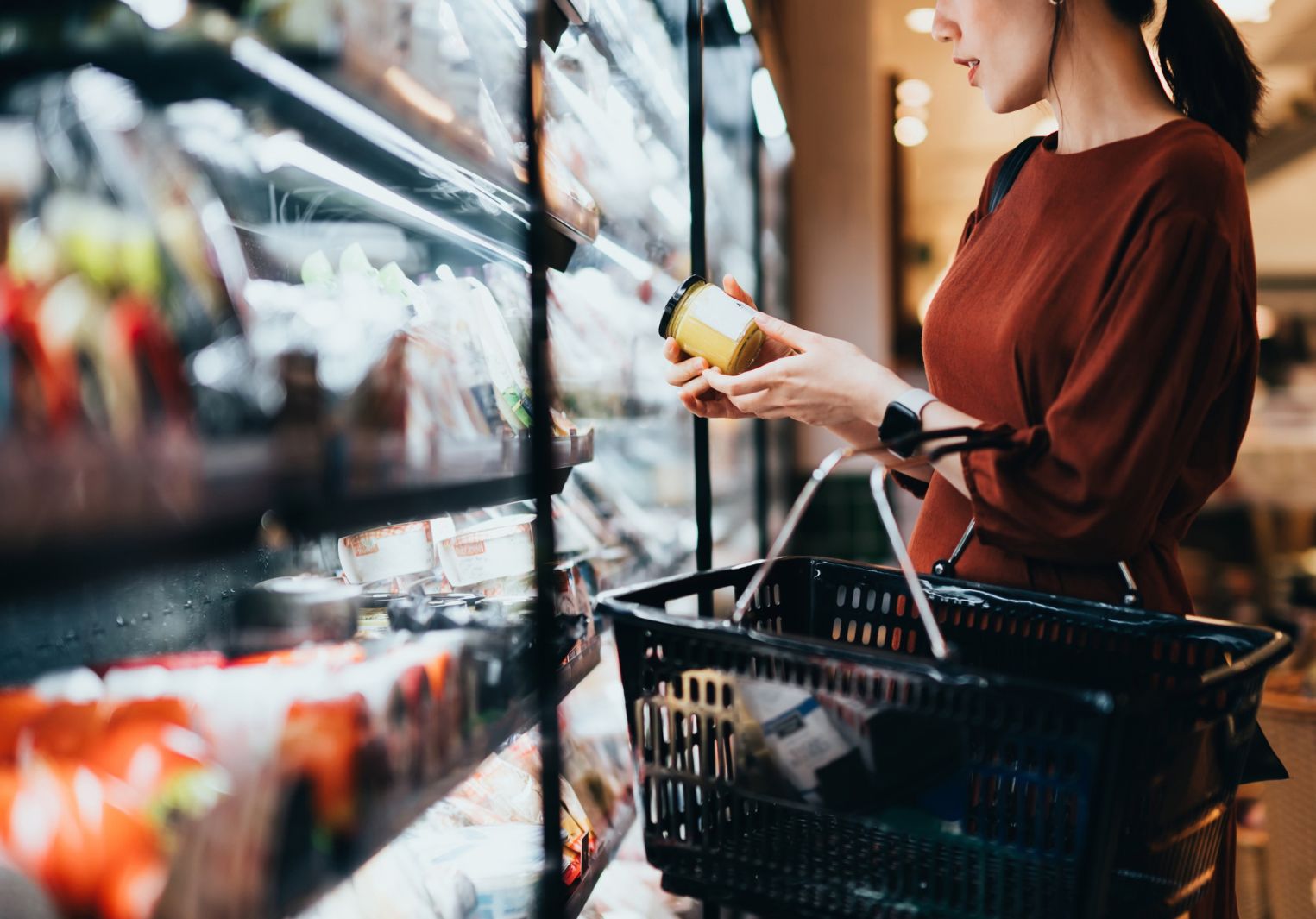 A young woman inspects a yellow jar in the middle of the fresh produce aisle, with a black basket hooked over her arm.