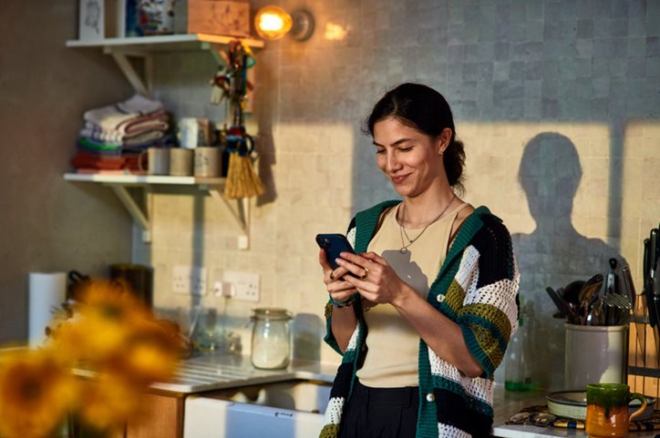 Woman stands in her kitchen looking at phone.