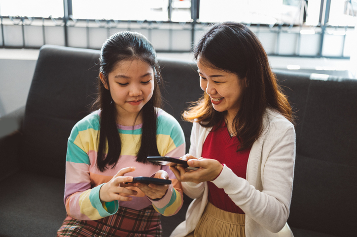 A young Asian teenage girl and her mother sitting side by side, both holding a smartphone and smiling.
