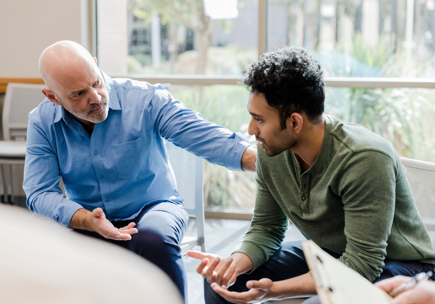 An older man speaking to a younger man with a hand on his shoulder. They are sitting side by side.
