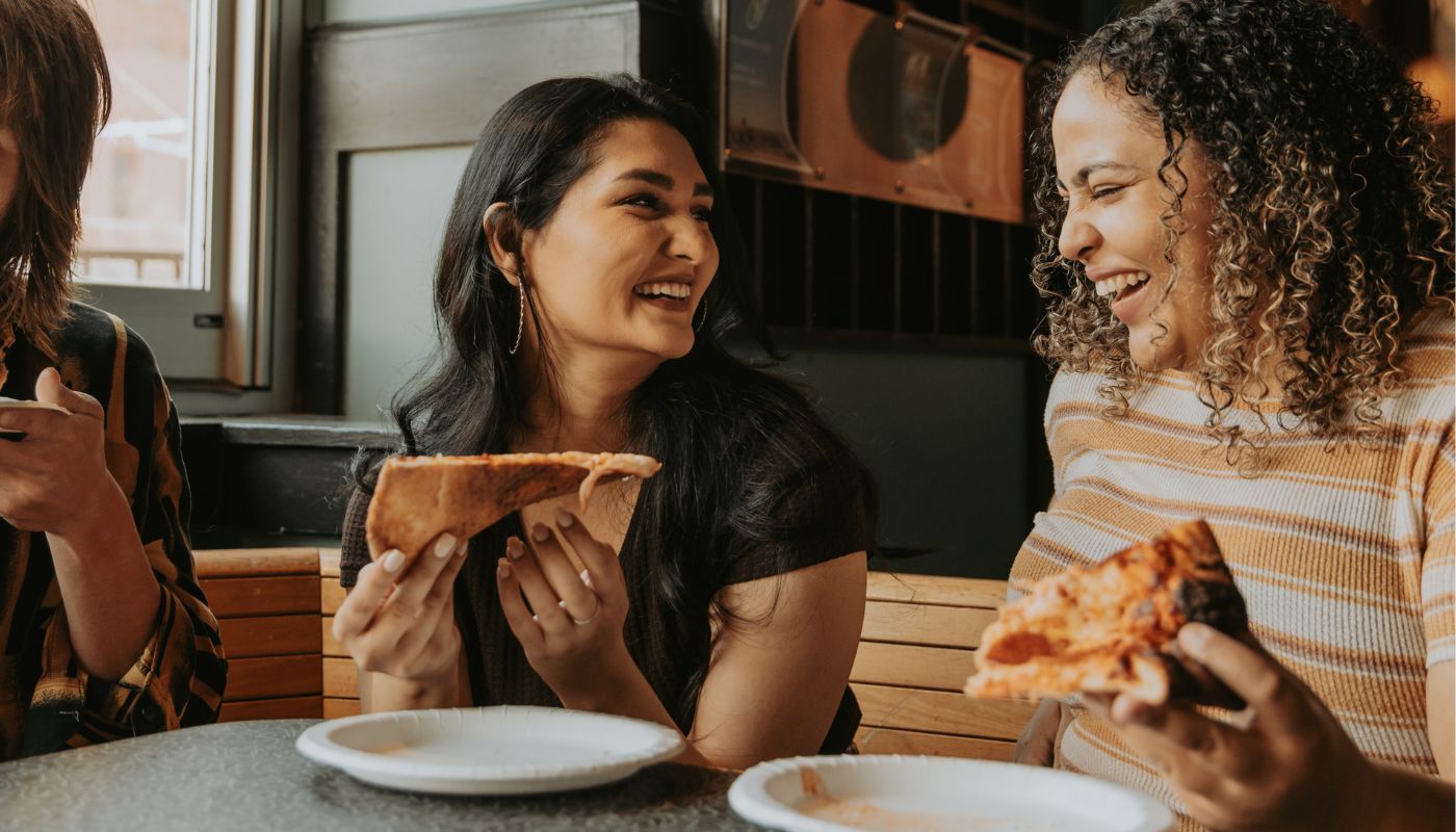 Two friends laugh and talk as they eat pizza.