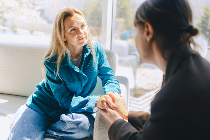 Two middle aged women holding hands over the arm of a couch and looking at each other in support.